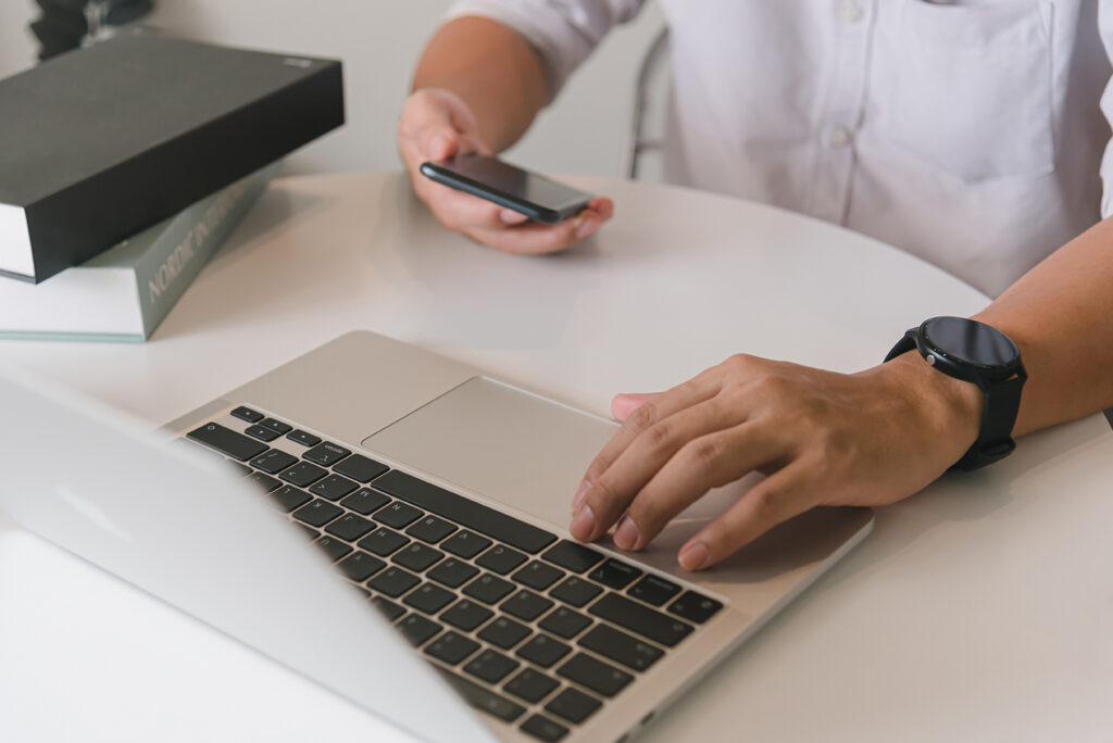 Mann sitzt Zuhause an einem Tisch und bedient einen Laptop und ein Smartphone.